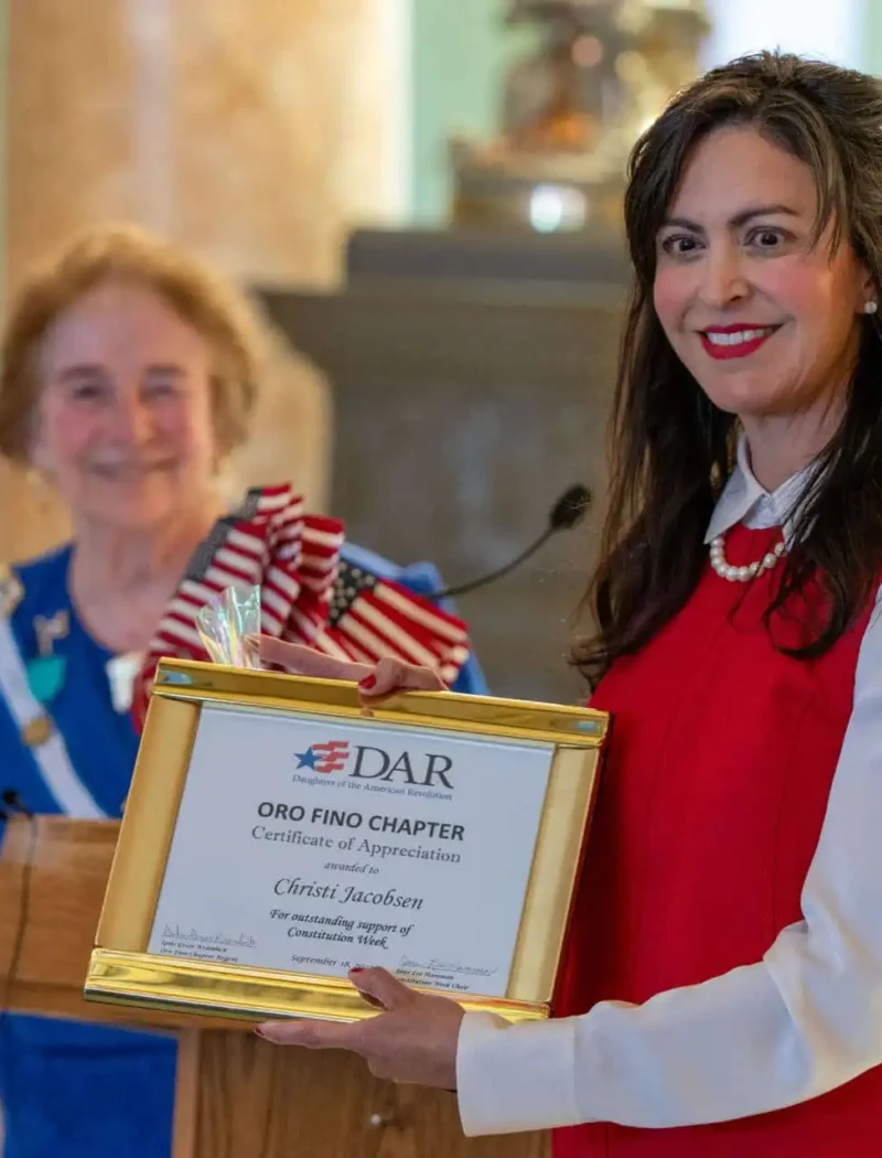 Secretary Christi Jacobsen joins Constitution Day celebration at Montana State Capitol 2 A smiling woman in a red dress holds a framed certificate, standing beside an older woman in a blue dress with a sash. Both are at a podium indoors, with another person blurred in the background.