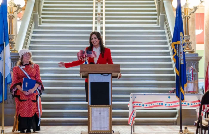 Montana Secretary of State Christi Jacobsen celebrates Constitution Day, announces 2022 Constitution Contest for Montana students K-12 1 A woman in a red blazer speaks at a podium decorated with small American flags during a Constitution Contest event, while another woman in colonial attire stands nearby. Patriotic decorations and a staircase complete the Constitution Day scene.