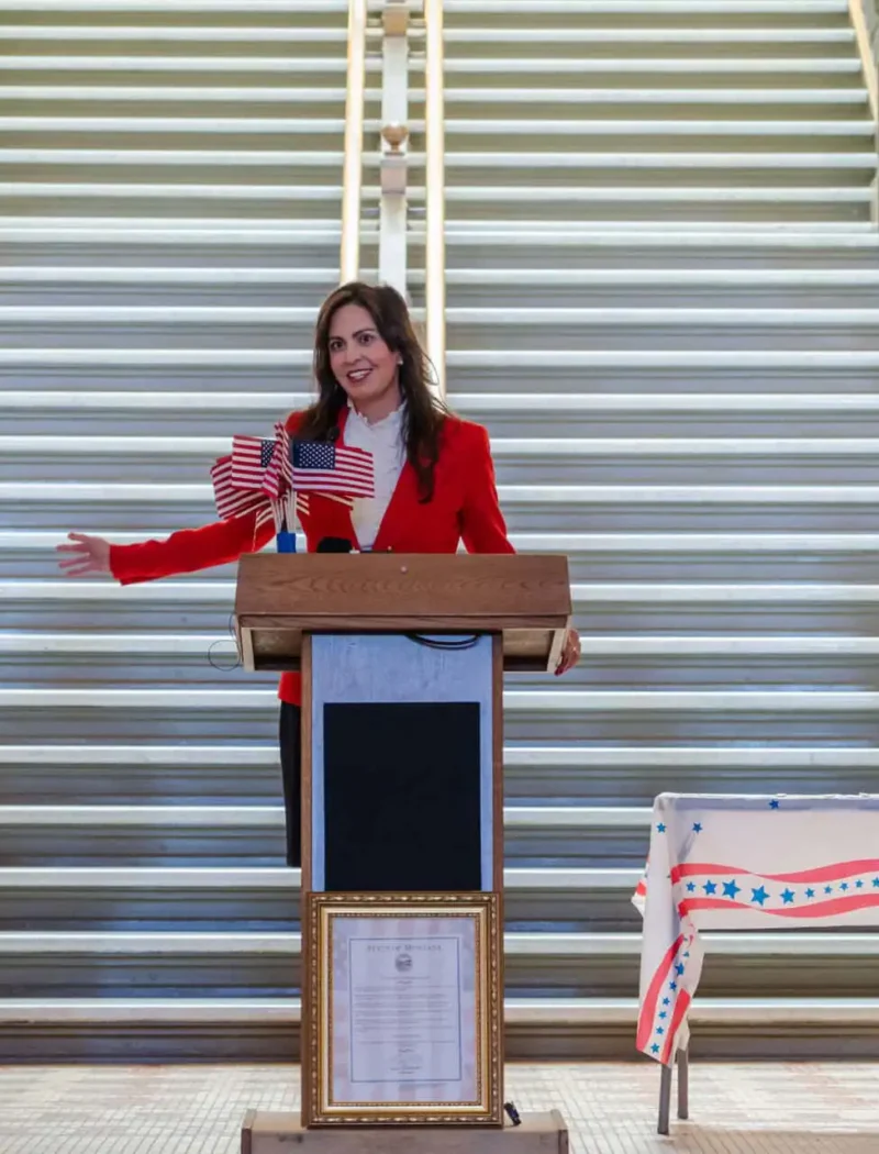 Montana Secretary of State Christi Jacobsen celebrates Constitution Day, announces 2022 Constitution Contest for Montana students K-12 3 A woman in a red blazer speaks at a podium decorated with small American flags during a Constitution Contest event, while another woman in colonial attire stands nearby. Patriotic decorations and a staircase complete the Constitution Day scene.