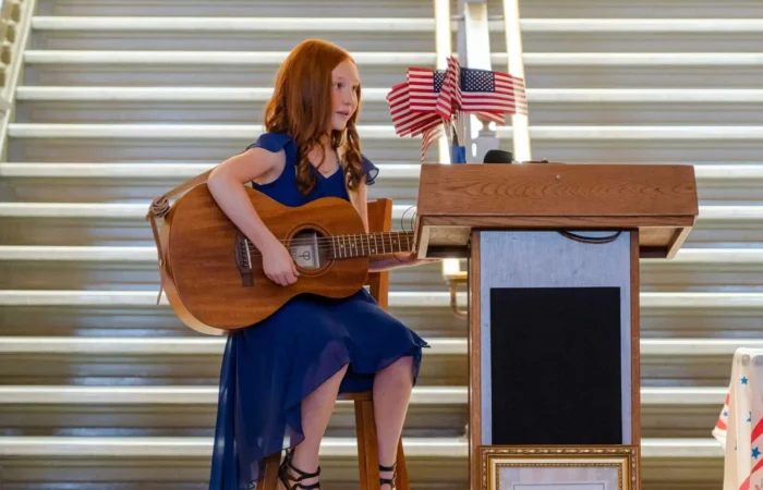 2021 US Constitution Contest winner Molly Gilbert performs at Montana State Capitol 9 A young Montana student in a blue dress sits on a stool, playing an acoustic guitar next to a wooden podium decorated with small American flags—possibly at a Constitution Contest hosted by the Montana Secretary of State; stairs are visible behind her.