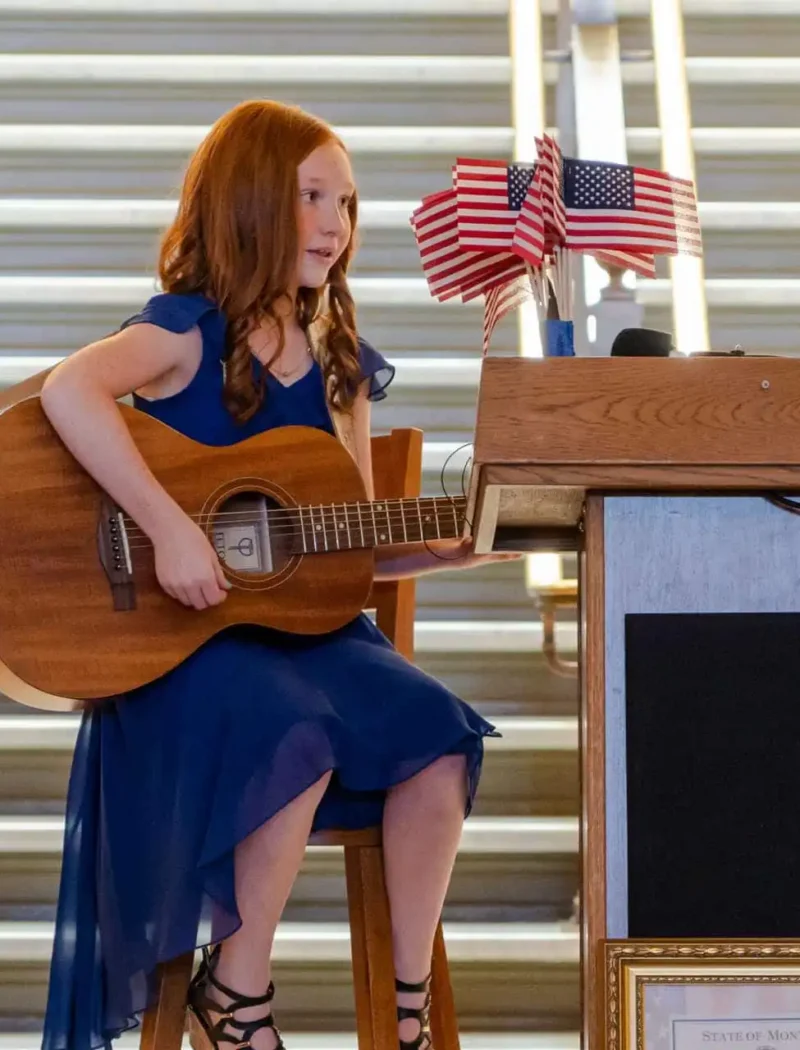 2021 US Constitution Contest winner Molly Gilbert performs at Montana State Capitol 1 A young Montana student in a blue dress sits on a stool, playing an acoustic guitar next to a wooden podium decorated with small American flags—possibly at a Constitution Contest hosted by the Montana Secretary of State; stairs are visible behind her.