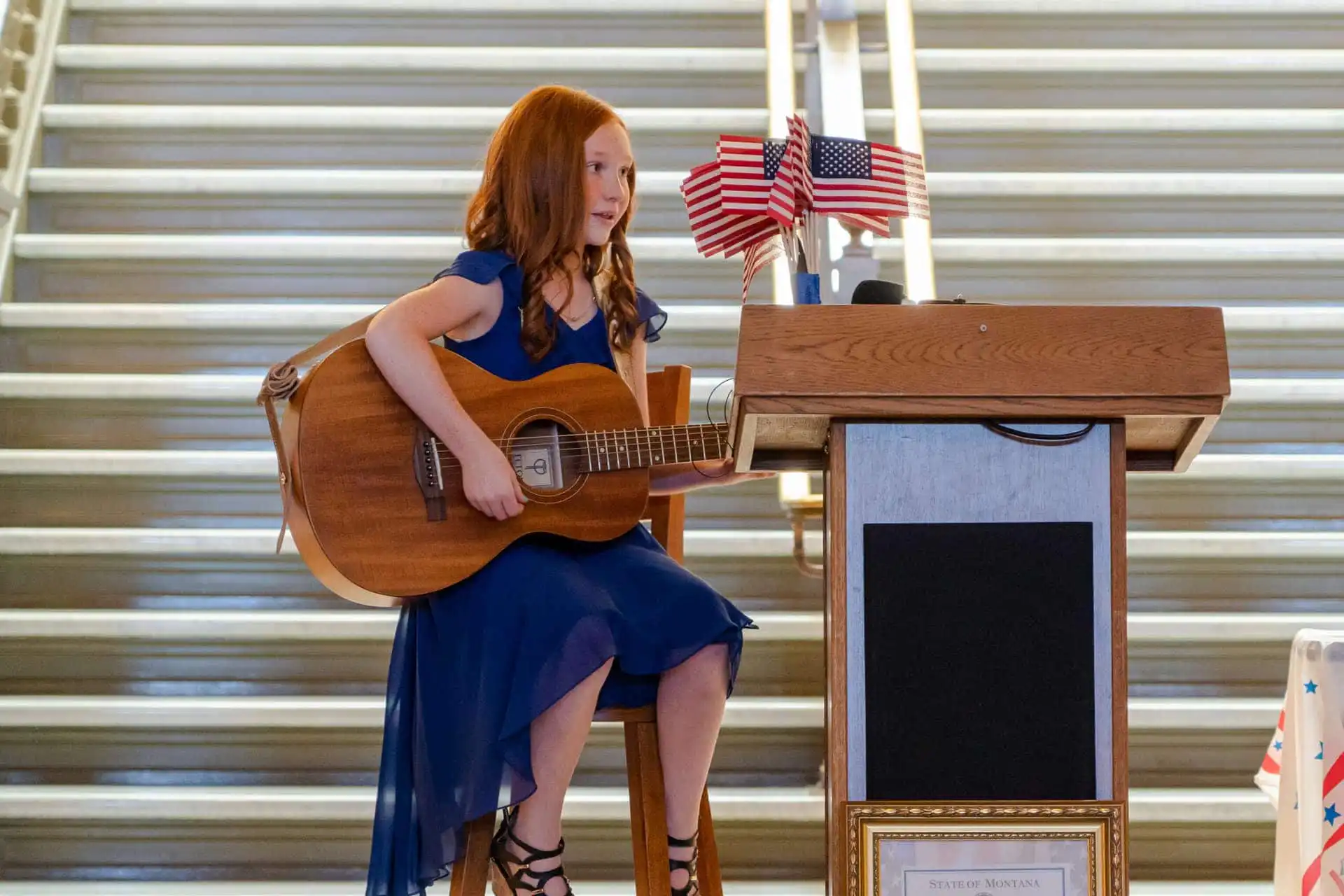 2021 US Constitution Contest winner Molly Gilbert performs at Montana State Capitol 2 A young Montana student in a blue dress sits on a stool, playing an acoustic guitar next to a wooden podium decorated with small American flags—possibly at a Constitution Contest hosted by the Montana Secretary of State; stairs are visible behind her.