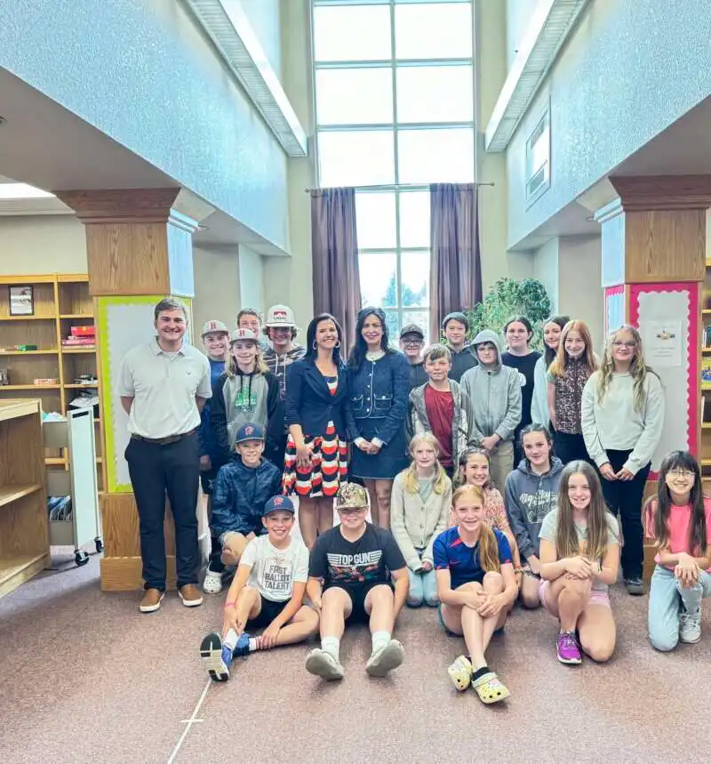 East Helena students and a few adults pose for a photo inside a library, with bookshelves and large windows in the background. Some people are standing, while others are seated on the floor.