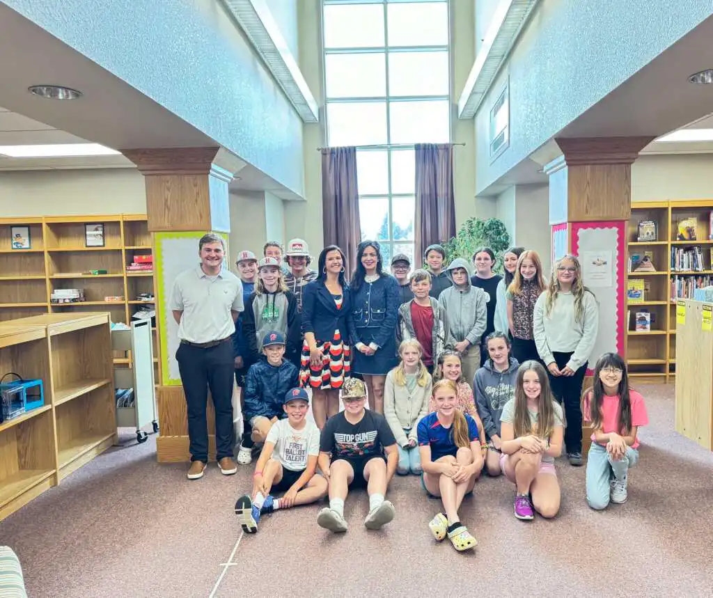 East Helena students and a few adults pose for a photo inside a library, with bookshelves and large windows in the background. Some people are standing, while others are seated on the floor.