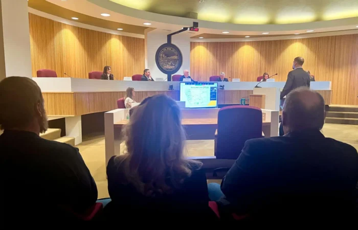 People sit in an audience while a person stands and presents information on a screen to the Montana Land Board, including Christi Jacobsen, and other officials seated at a long desk in a modern chamber discussing Montana revenue.