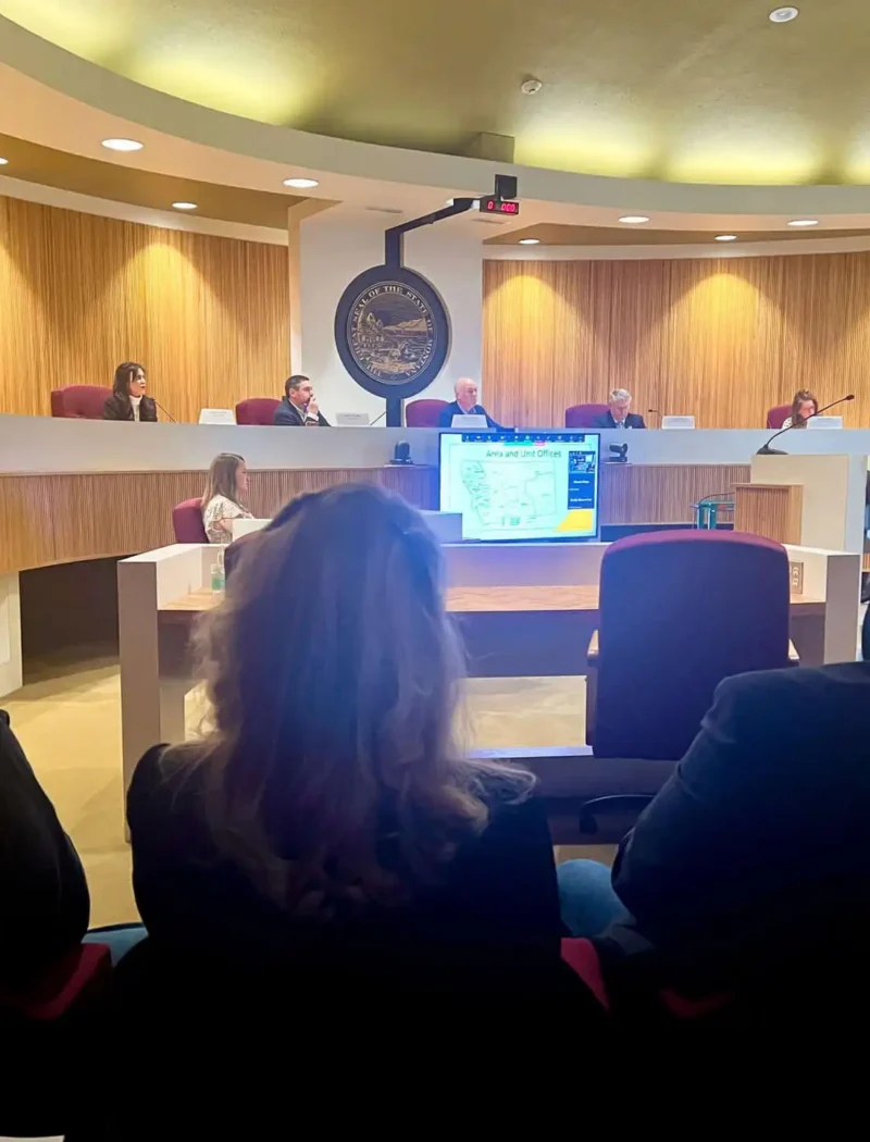People sit in an audience while a person stands and presents information on a screen to the Montana Land Board, including Christi Jacobsen, and other officials seated at a long desk in a modern chamber discussing Montana revenue.