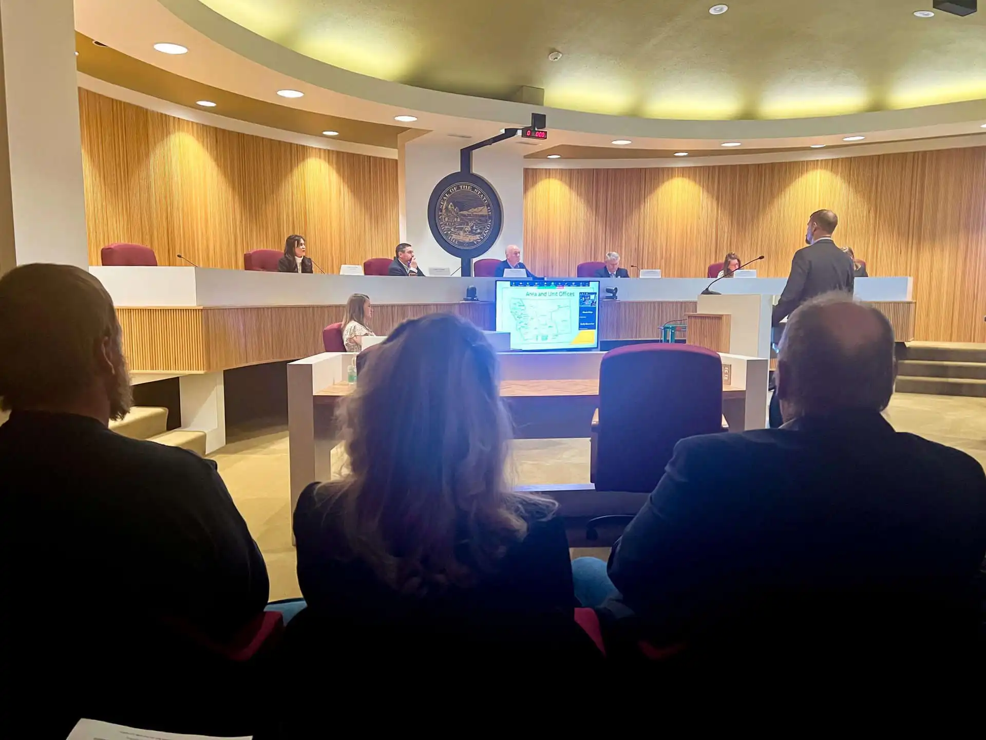 People sit in an audience while a person stands and presents information on a screen to the Montana Land Board, including Christi Jacobsen, and other officials seated at a long desk in a modern chamber discussing Montana revenue.