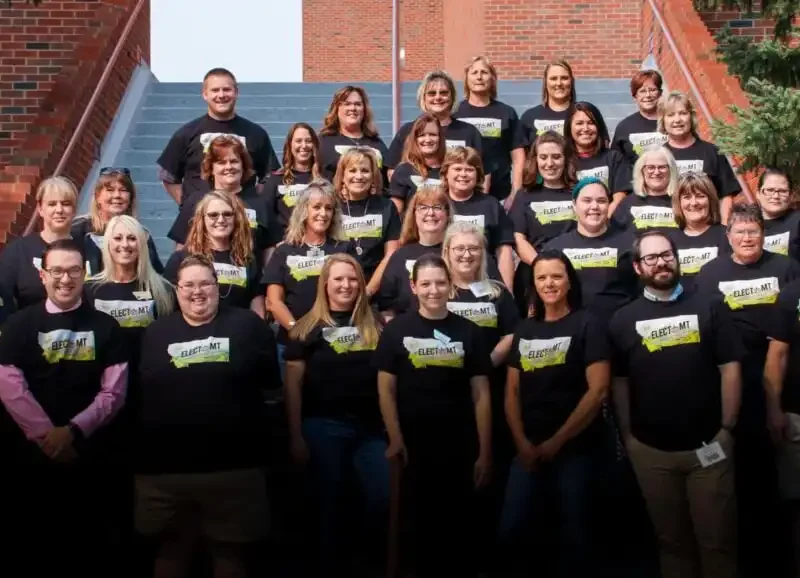 A group of adults, wearing matching black T-shirts with a yellow and white graphic, pose for a group photo on outdoor steps surrounded by brick walls and greenery during electMT training organized by the Elections Division of the Secretary of State.