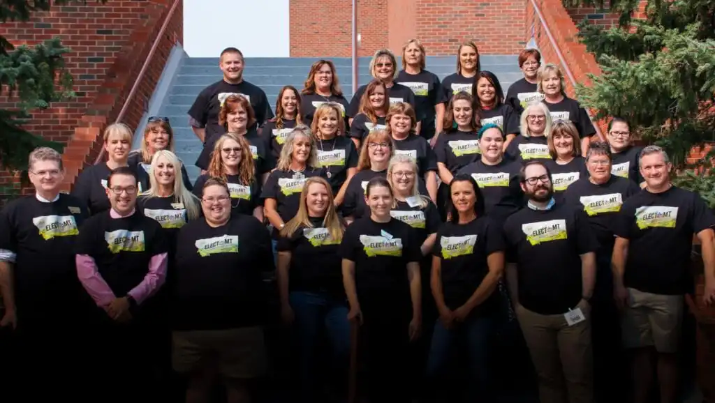Secretary of State's Elections Division begins electMT training 14 A group of adults, wearing matching black T-shirts with a yellow and white graphic, pose for a group photo on outdoor steps surrounded by brick walls and greenery during electMT training organized by the Elections Division of the Secretary of State.