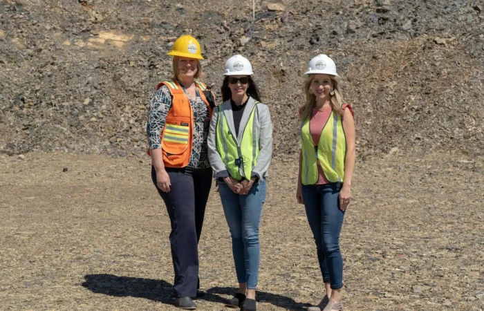 Secretary Christi Jacobsen given tour of Black Butte Copper Project 7 Three women stand outdoors on rocky ground at the Black Butte Copper Project, wearing safety gear including hard hats and reflective vests. Two wear yellow, one wears orange, and all are smiling at the camera. Christi Jacobsen is among them.