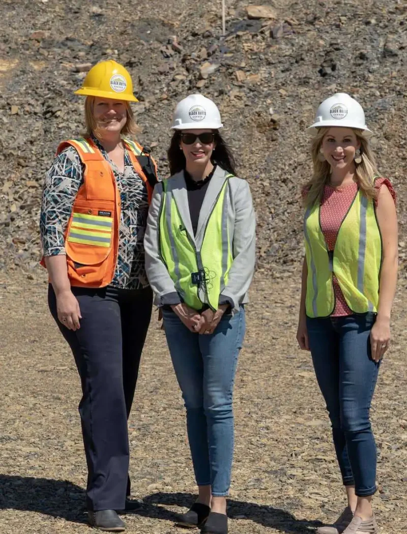 Three women stand outdoors on rocky ground at the Black Butte Copper Project, wearing safety gear including hard hats and reflective vests. Two wear yellow, one wears orange, and all are smiling at the camera. Christi Jacobsen is among them.