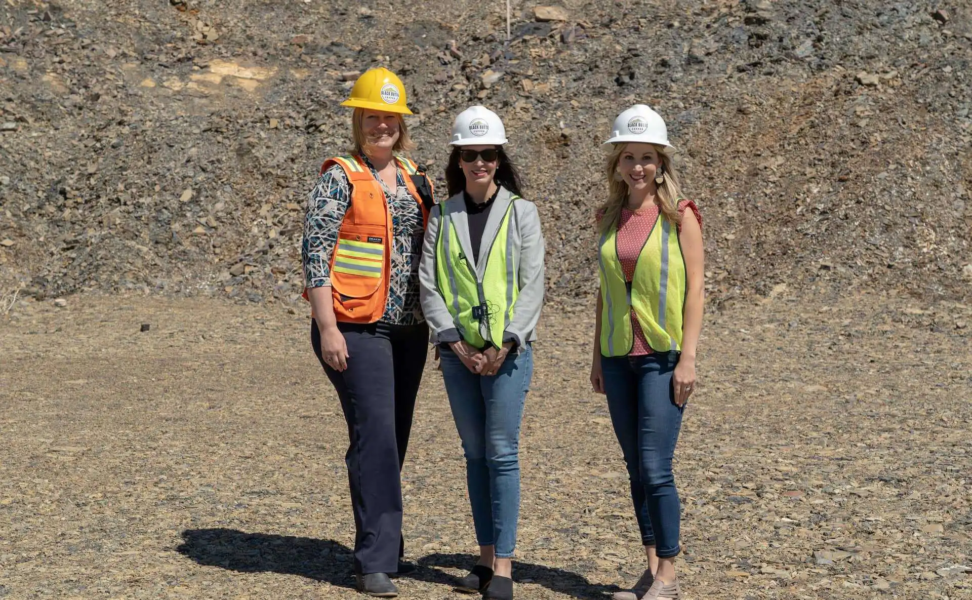 Three women stand outdoors on rocky ground at the Black Butte Copper Project, wearing safety gear including hard hats and reflective vests. Two wear yellow, one wears orange, and all are smiling at the camera. Christi Jacobsen is among them.