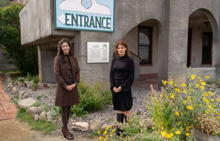 Montana Business Spotlight: Ghosts and geothermals part of the history at Boulder Hot Springs 3 Two women stand outside Boulder Hot Springs near a large Entrance sign and a garden with yellow flowers. The geothermal retreat in Montana features gray walls, arched windows, and a plaque mounted beside the door.