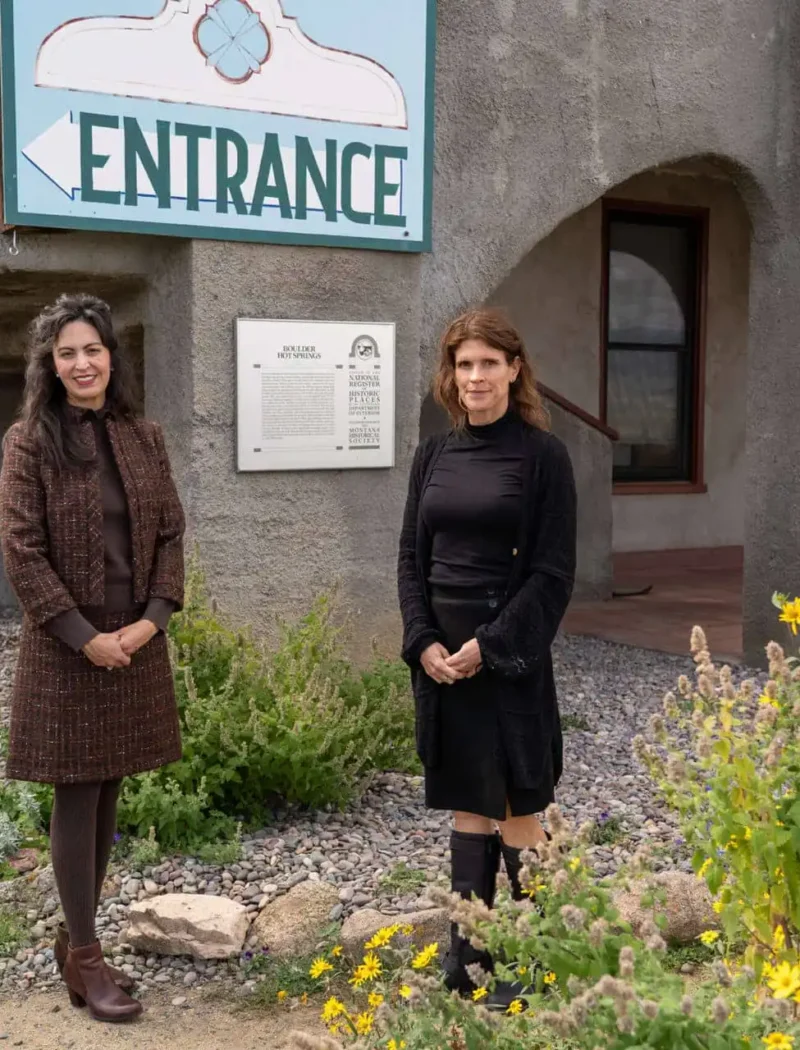 Two women stand outside Boulder Hot Springs near a large Entrance sign and a garden with yellow flowers. The geothermal retreat in Montana features gray walls, arched windows, and a plaque mounted beside the door.
