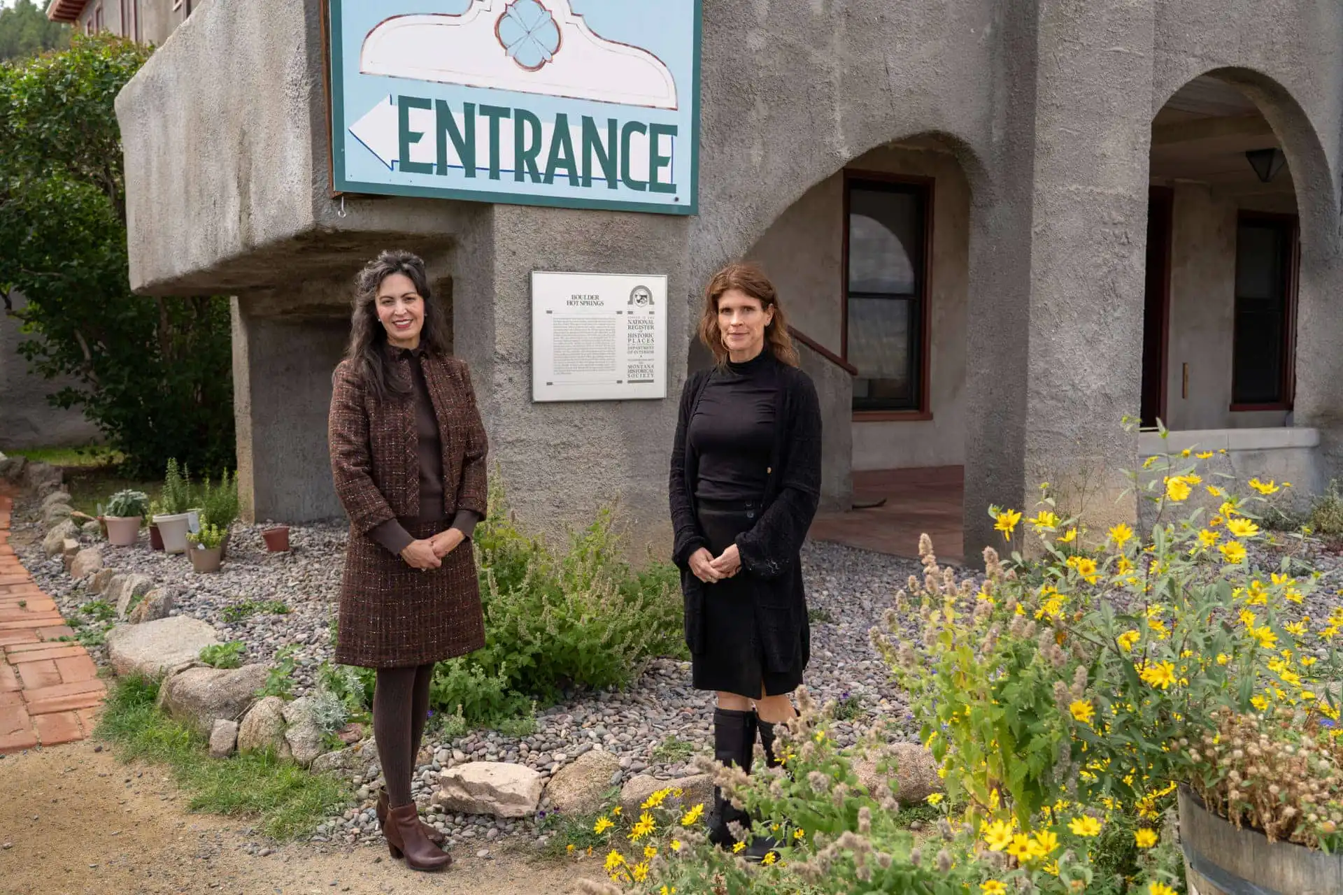 Two women stand outside Boulder Hot Springs near a large Entrance sign and a garden with yellow flowers. The geothermal retreat in Montana features gray walls, arched windows, and a plaque mounted beside the door.