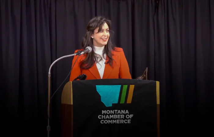 Secretary Jacobsen, in an orange blazer, speaks at a podium draped with the Montana Chamber of Commerce logo during Business Days at the Capitol. Black curtains frame the scene as she addresses topics like business registrations.