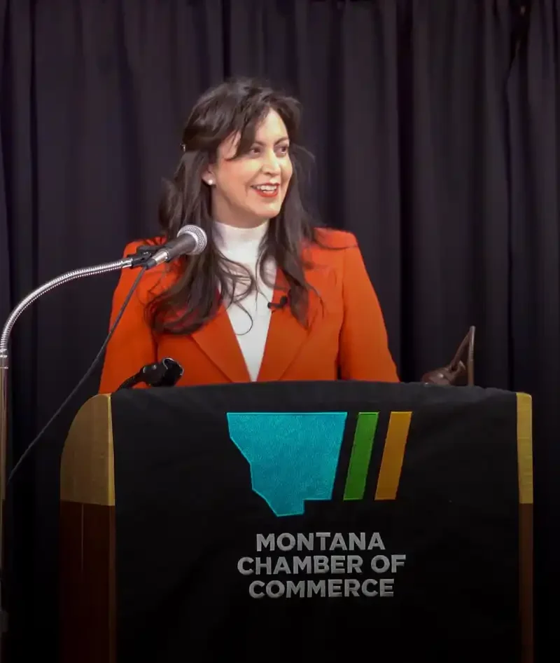 Secretary Jacobsen, in an orange blazer, speaks at a podium draped with the Montana Chamber of Commerce logo during Business Days at the Capitol. Black curtains frame the scene as she addresses topics like business registrations.