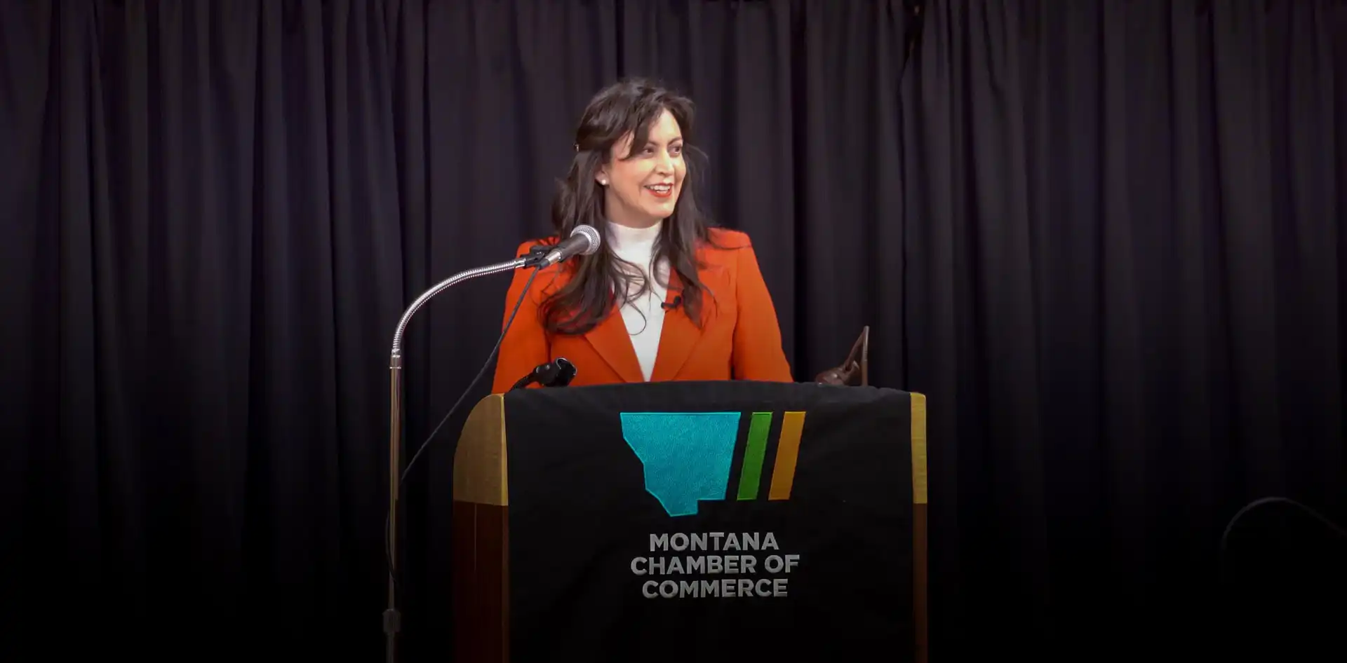 Secretary Jacobsen, in an orange blazer, speaks at a podium draped with the Montana Chamber of Commerce logo during Business Days at the Capitol. Black curtains frame the scene as she addresses topics like business registrations.