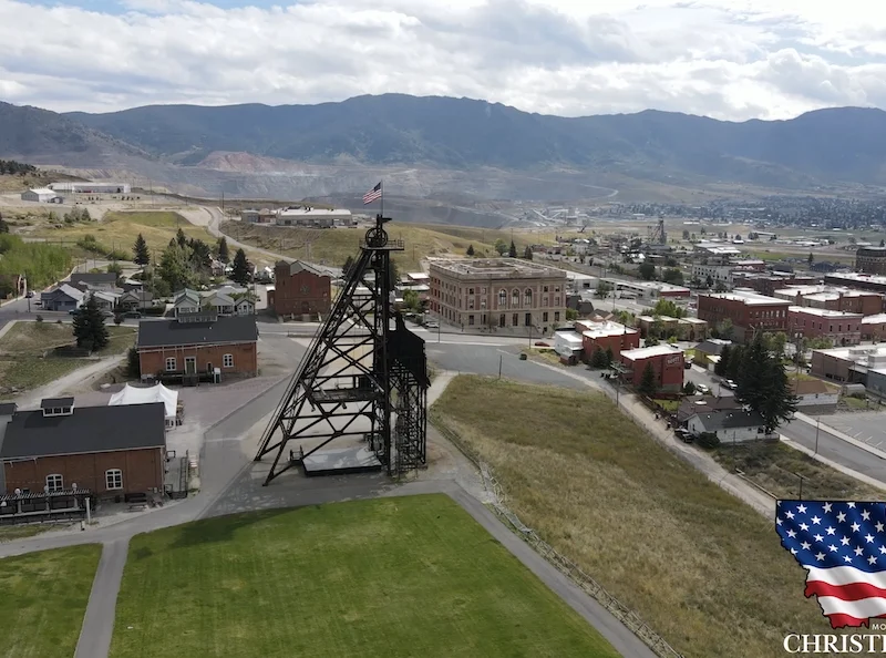 Aerial view of historic mining town in Butte with old mine structures, brick buildings, open grassy areas, and mountains in the background. An American flag, America250, and Secretary of State Christi Jacobsen appear in the bottom right corner.