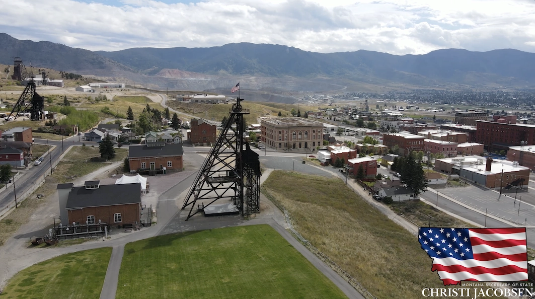 Aerial view of historic mining town in Butte with old mine structures, brick buildings, open grassy areas, and mountains in the background. An American flag, America250, and Secretary of State Christi Jacobsen appear in the bottom right corner.
