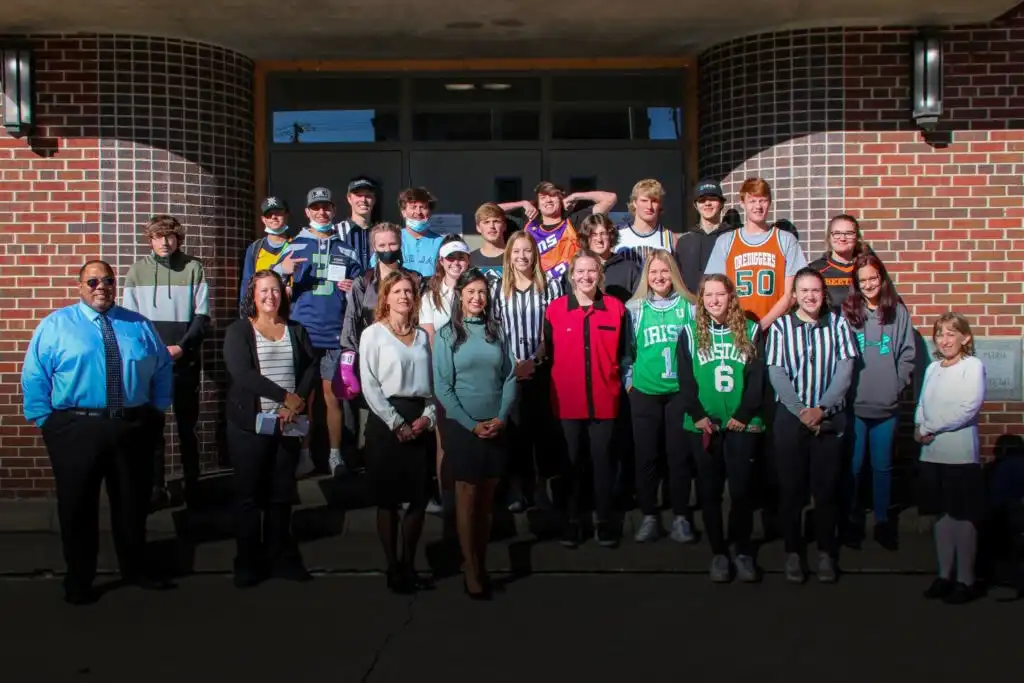 A group of Butte Central students and adults pose together outside a brick building. Some are wearing sports jerseys or referee shirts, and all are smiling in the sunlight.