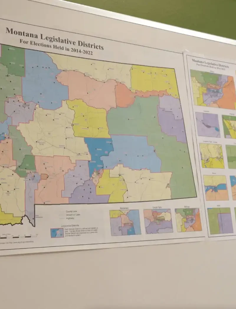 A whiteboard displays two posters with colorful maps of Montana legislative districts, highlighting various regions and districts, including a large detailed map and smaller inset maps on the right, relevant to election and 2022 candidate filing.