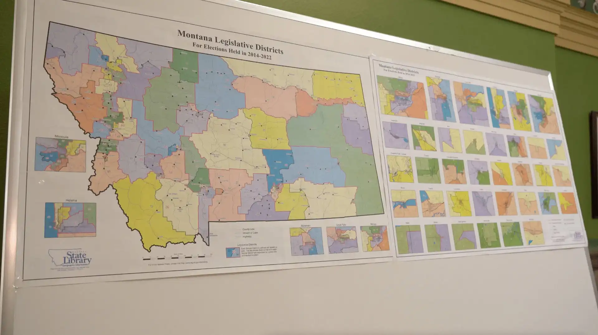 A whiteboard displays two posters with colorful maps of Montana legislative districts, highlighting various regions and districts, including a large detailed map and smaller inset maps on the right, relevant to election and 2022 candidate filing.
