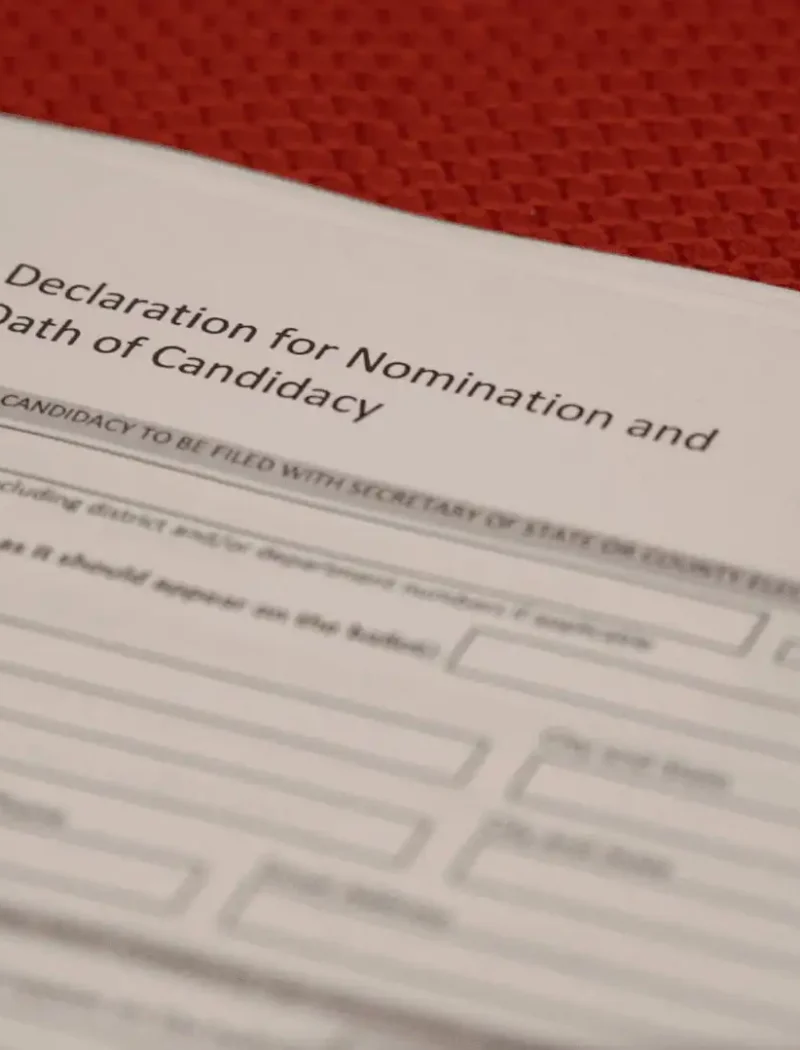 A close-up of a “Declaration for Nomination and Oath of Candidacy” form on a red textured surface, showing blanks for candidate information and the Montana state seal—a key document for 2022 candidate filing in the election.