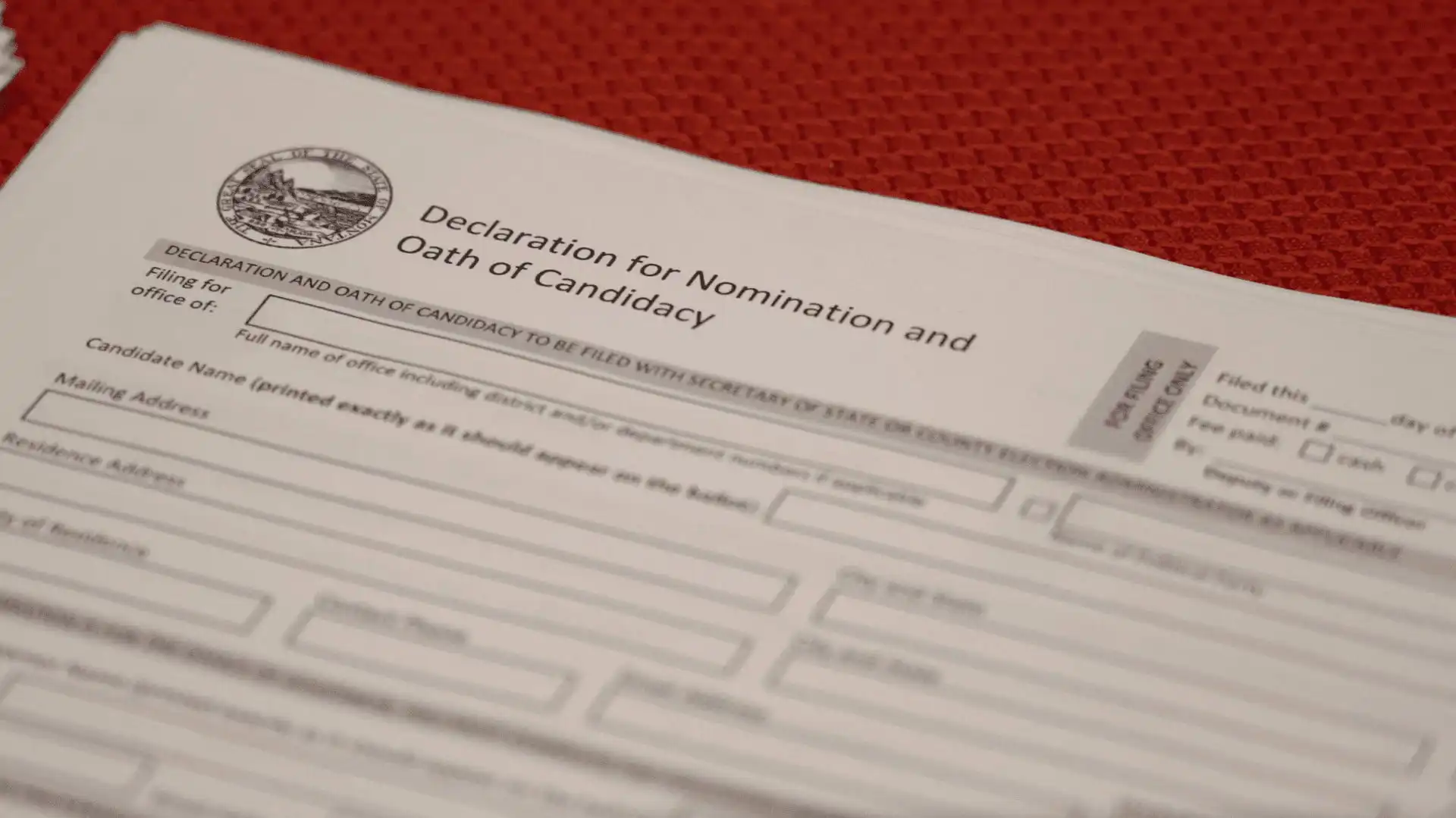 A close-up of a “Declaration for Nomination and Oath of Candidacy” form on a red textured surface, showing blanks for candidate information and the Montana state seal—a key document for 2022 candidate filing in the election.