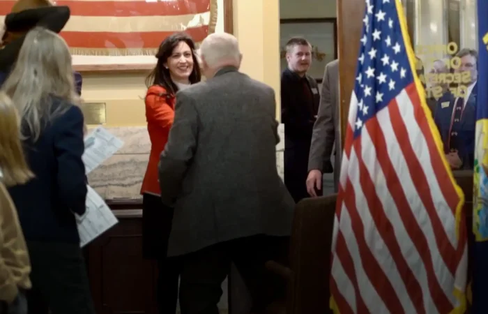 Eighty-one candidates file on first day of Montana's 2022 candidate filing 3 A woman in a red jacket smiles and shakes hands with an older man in a gray jacket in an office with American flags and people standing around, some holding papers—likely Montana candidates during 2022 candidate filing. An American flag is in the foreground.