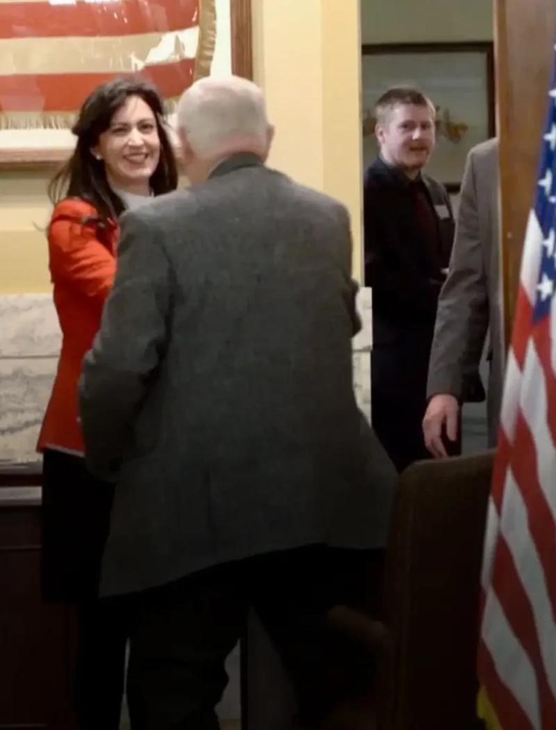 A woman in a red jacket smiles and shakes hands with an older man in a gray jacket in an office with American flags and people standing around, some holding papers—likely Montana candidates during 2022 candidate filing. An American flag is in the foreground.