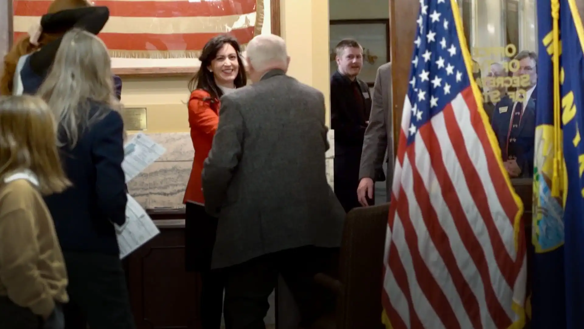 A woman in a red jacket smiles and shakes hands with an older man in a gray jacket in an office with American flags and people standing around, some holding papers—likely Montana candidates during 2022 candidate filing. An American flag is in the foreground.
