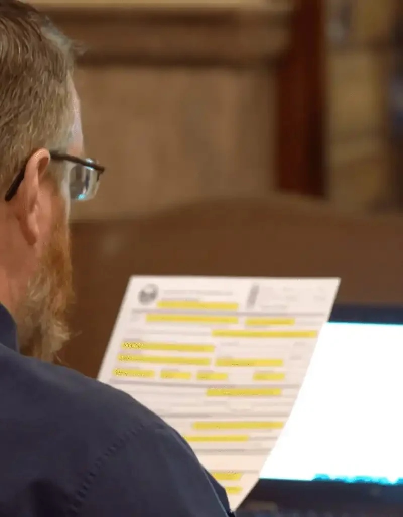 A man with glasses and a beard sits at a desk, reading highlighted candidate filings. An open laptop glows in front of him as he reviews new candidate forms, likely related to the Secretary of State, in an indoor formal setting.