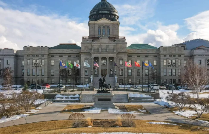 The Montana State Capitol building in Helena, with a domed roof, state and U.S. flags in front, is where the Montana Secretary of State oversees elections and law enforcement helps prevent voting crime, all framed by snow patches, trees, and distant hills under a partly cloudy sky.