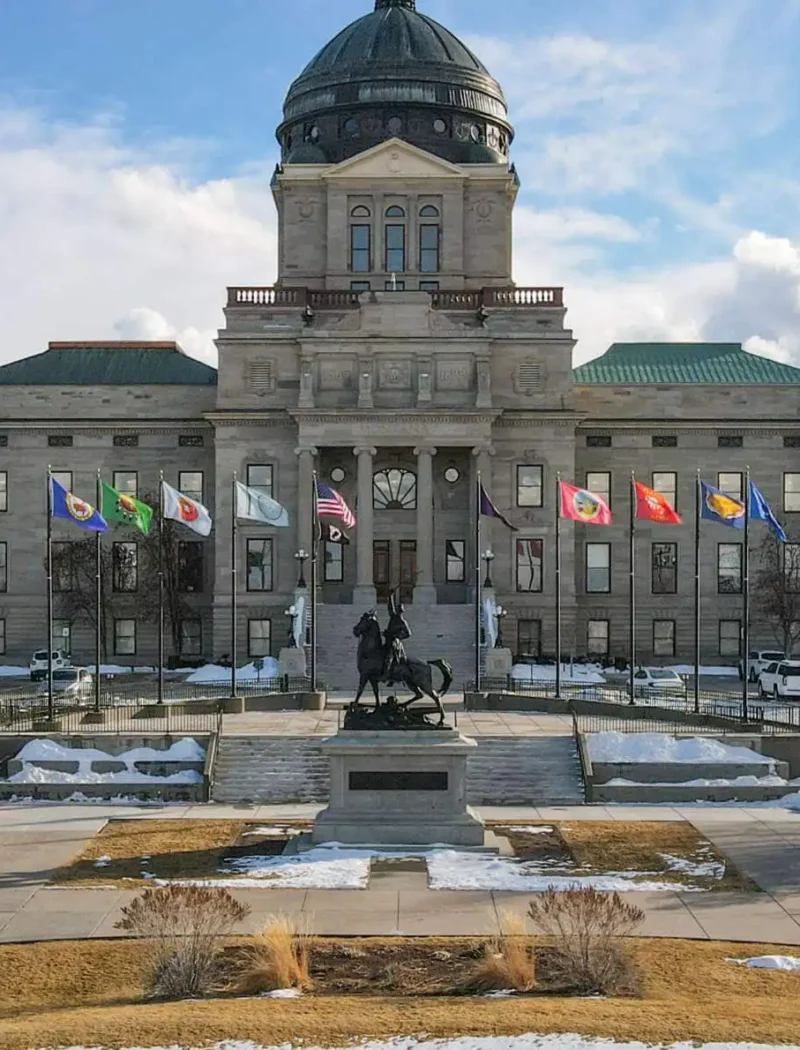 The Montana State Capitol building in Helena, with a domed roof, state and U.S. flags in front, is where the Montana Secretary of State oversees elections and law enforcement helps prevent voting crime, all framed by snow patches, trees, and distant hills under a partly cloudy sky.