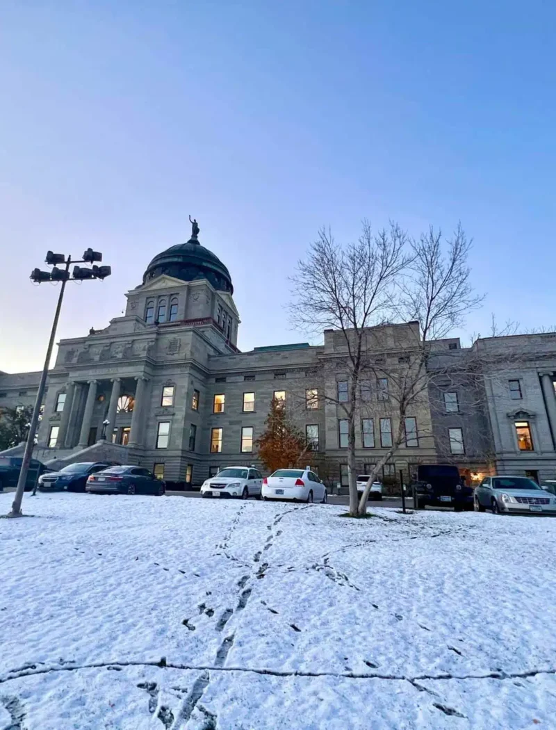 A historic stone building with a central dome stands under a clear sky, surrounded by parked cars and snow-covered ground crisscrossed with footprints. Leafless trees line the front—an iconic scene during Montana precincts election results tally.
