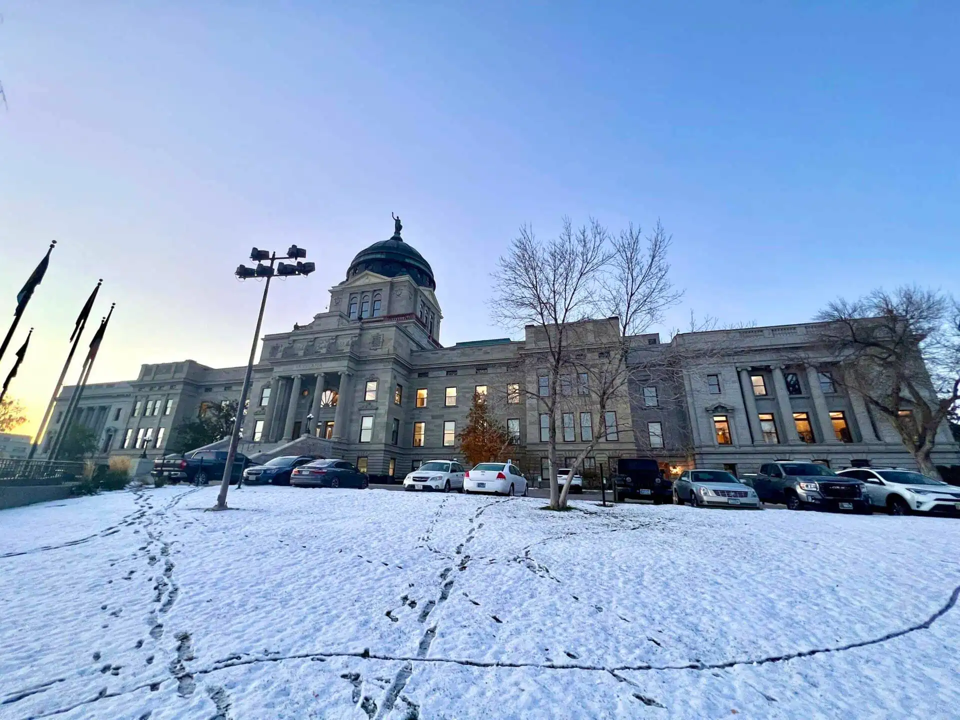 A historic stone building with a central dome stands under a clear sky, surrounded by parked cars and snow-covered ground crisscrossed with footprints. Leafless trees line the front—an iconic scene during Montana precincts election results tally.