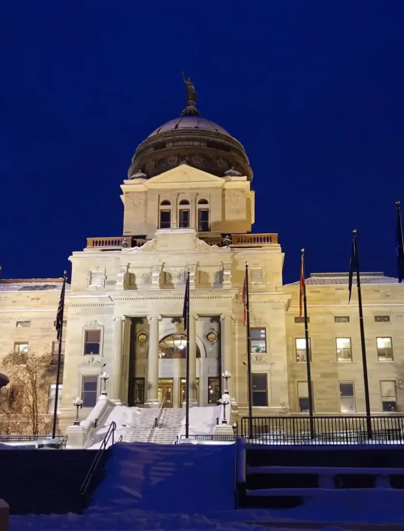 The illuminated Montana State Capitol building is seen at night with snow on the ground. A bronze equestrian statue stands in the foreground, and several flags are displayed—symbolizing Montana’s commitment to implement a secure election management system.