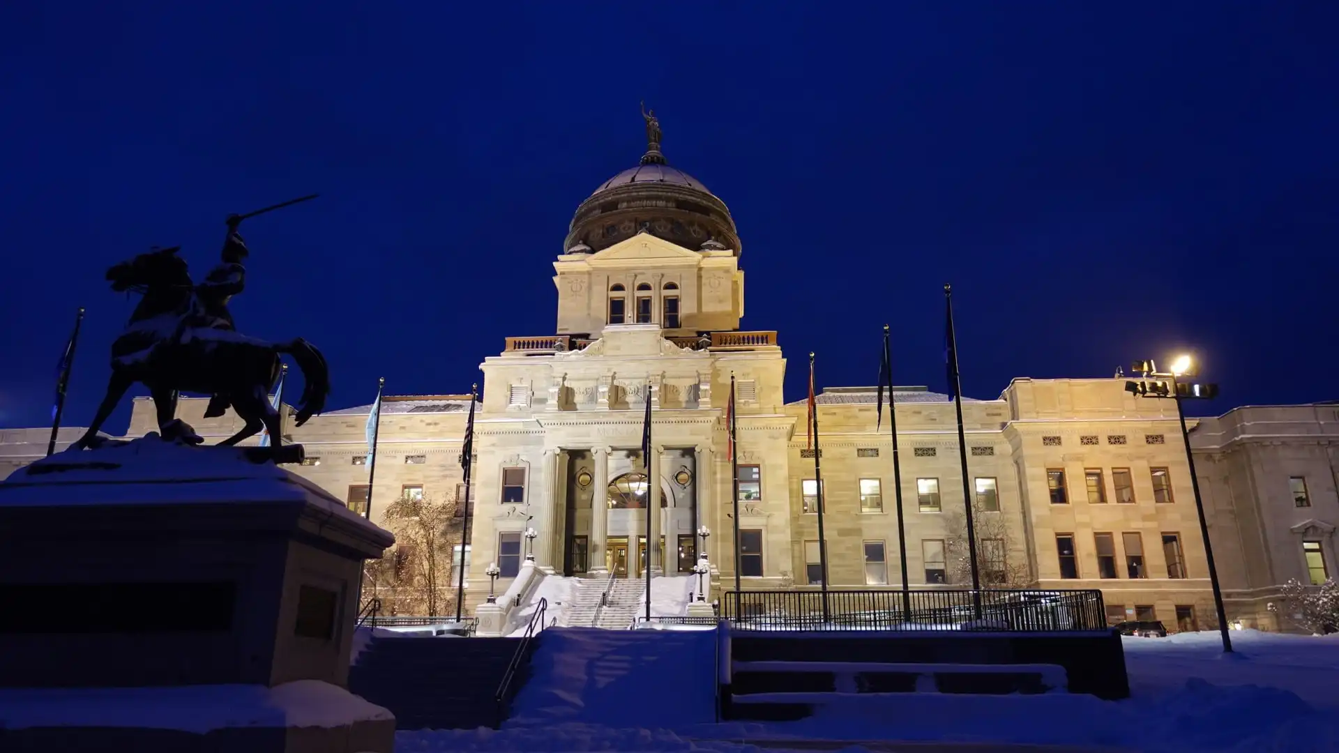 Montana State Capitol building at night, illuminated with lights, snow on the ground, and a statue of a rider on horseback in the foreground. Dark blue sky overhead.