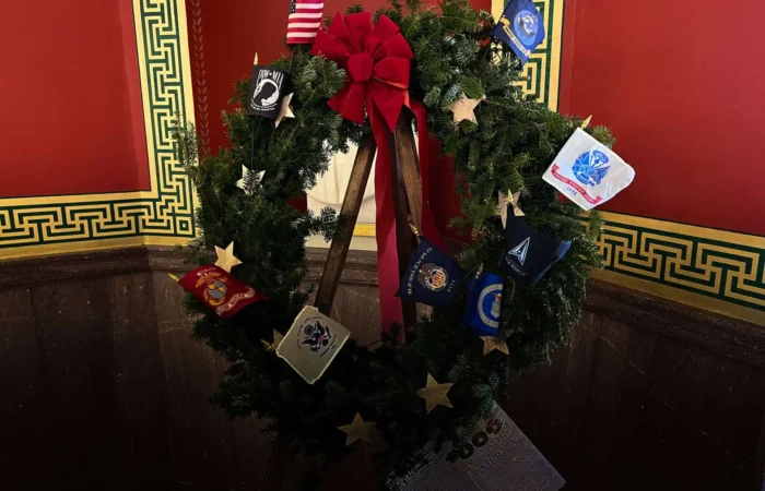 A holiday wreath with a red bow is decorated with military service emblems and gold stars, honoring Wreaths Across America, displayed against a red wall with ornate green and gold trim.