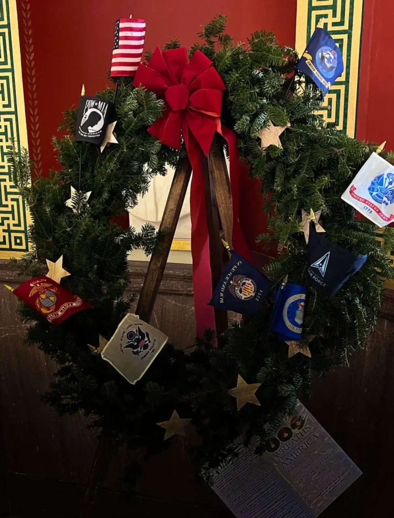 A holiday wreath with a red bow is decorated with military service emblems and gold stars, honoring Wreaths Across America, displayed against a red wall with ornate green and gold trim.