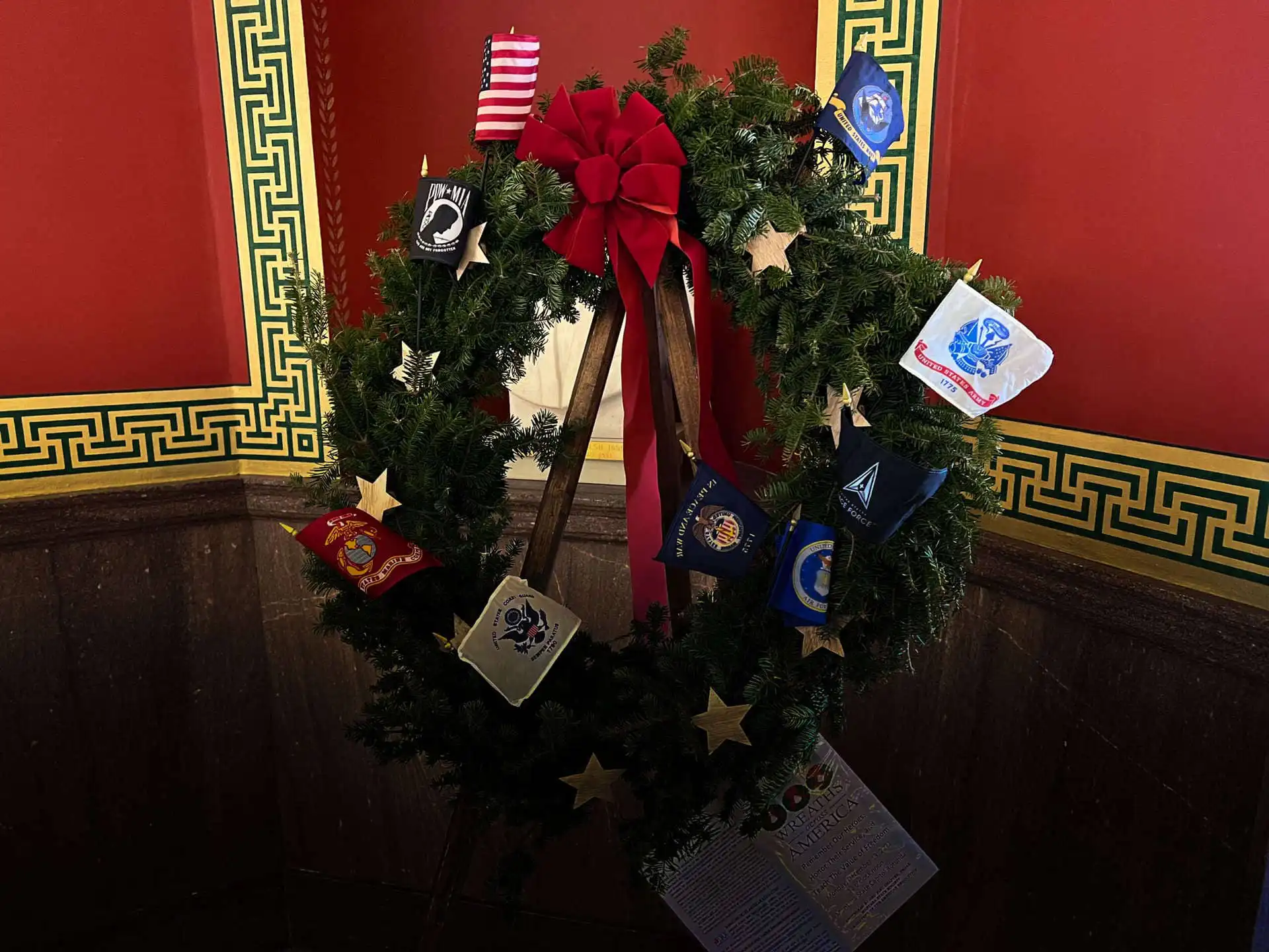 Secretary Jacobsen attends Wreaths Across America ceremony in capitol rotunda 8 A holiday wreath with a red bow is decorated with military service emblems and gold stars, honoring Wreaths Across America, displayed against a red wall with ornate green and gold trim.