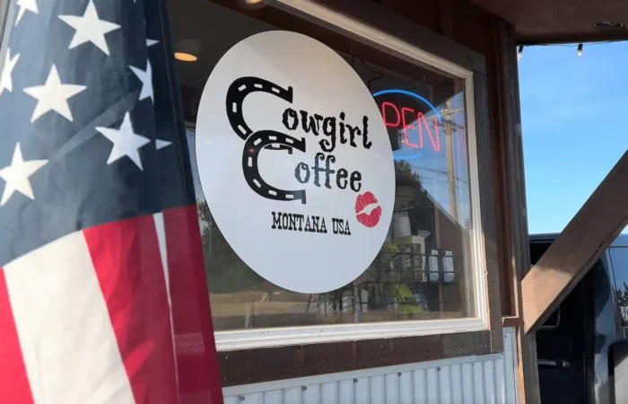 Montana Business Spotlight: Cowgirl Coffee 'super appreciative' nearing 20-year anniversary 9 A storefront window displays the Cowgirl Coffee Montana USA logo with a red lipstick kiss mark, celebrating 20-year anniversary. An American flag stands in the foreground, and a neon OPEN sign glows in this iconic Montana Business.