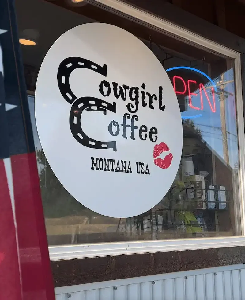 A storefront window displays the Cowgirl Coffee Montana USA logo with a red lipstick kiss mark, celebrating 20-year anniversary. An American flag stands in the foreground, and a neon OPEN sign glows in this iconic Montana Business.