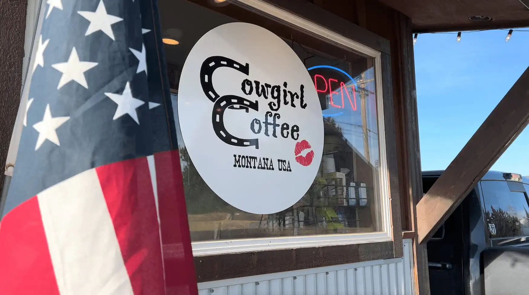 A storefront window displays the Cowgirl Coffee Montana USA logo with a red lipstick kiss mark, celebrating 20-year anniversary. An American flag stands in the foreground, and a neon OPEN sign glows in this iconic Montana Business.