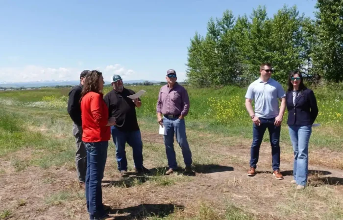 A group of six adults stand outdoors in a grassy field under a blue sky. Some are talking, while others face the camera. Green trees and wildflowers are visible in the background.