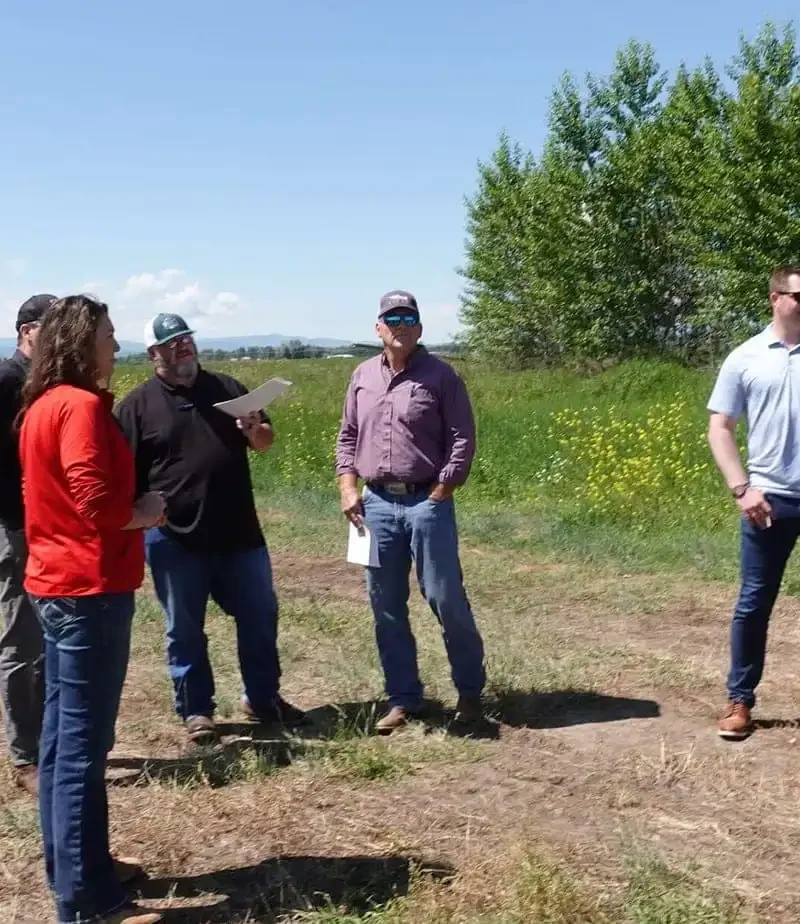 A group of six adults stand outdoors in a grassy field under a blue sky. Some are talking, while others face the camera. Green trees and wildflowers are visible in the background.