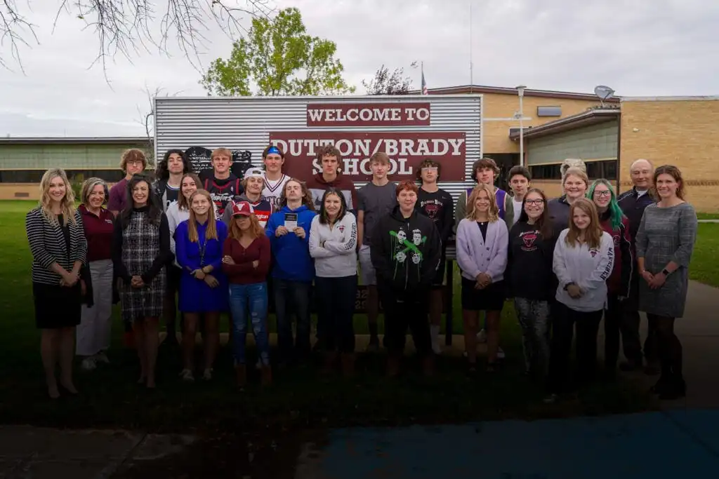 A group of students and adults pose together in front of Dutton-Brady Schools, where Secretary Jacobsen delivers Constitutions. They stand on grass under a cloudy sky by a sign that reads Welcome to Dutton/Brady School.
