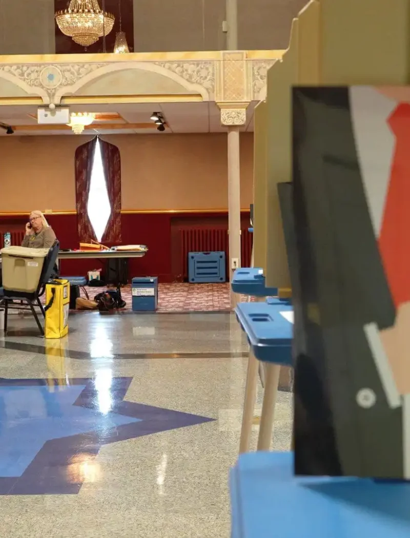 A voting center with poll workers seated at a table in the background and a sign in the foreground displaying voter information with a checkmark and a suit illustration, captured during Montana Election season. Blue voting booths line the right side of the image.