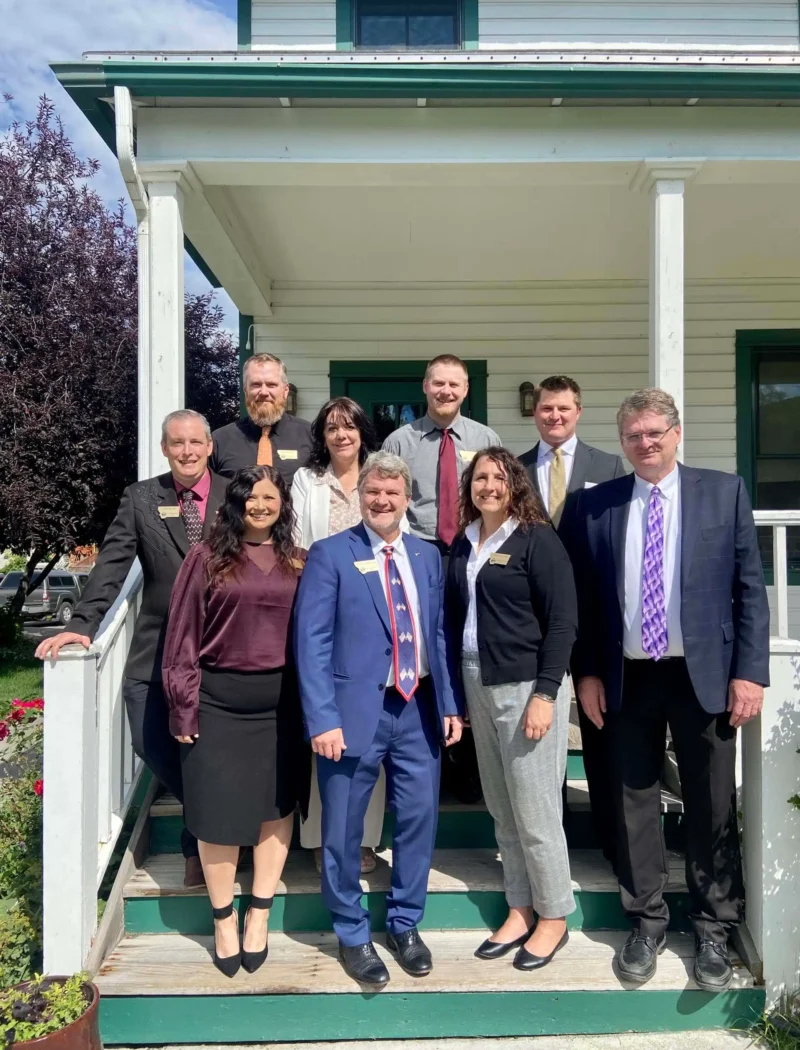 Secretary of State’s Office presents at 2023 Montana Association of Clerks & Recorders Convention 9 Nine adults in business attire, smiling and posing on the steps of a white house with green trim during the sunny 2023 Convention of the Montana Association of Clerks & Recorders. Trees and grass frame the group, which includes both men and women.